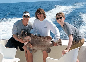 Three anglers sitting on transom holding a huge Grouper.